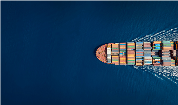 Overhead view of a container ship entering from the right side of the frame with a wake streaming behind it; open ocean fills the rest of the image.