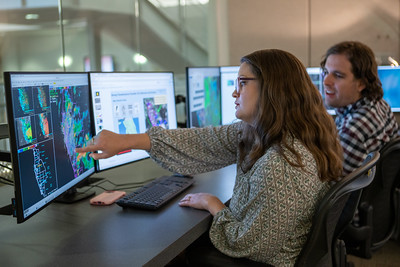 A woman is seated at a desk, pointing to a radar map on a computer screen; her male coworker looks on, smiling. Additional computer screens in front of them feature presentation slides, maps, and spreadsheets.