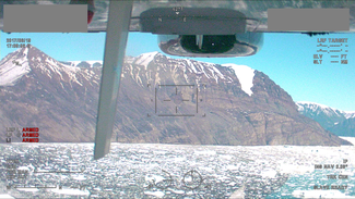 Image from the inside of an airplane showing a steep mountainside by water partially covered in ice.