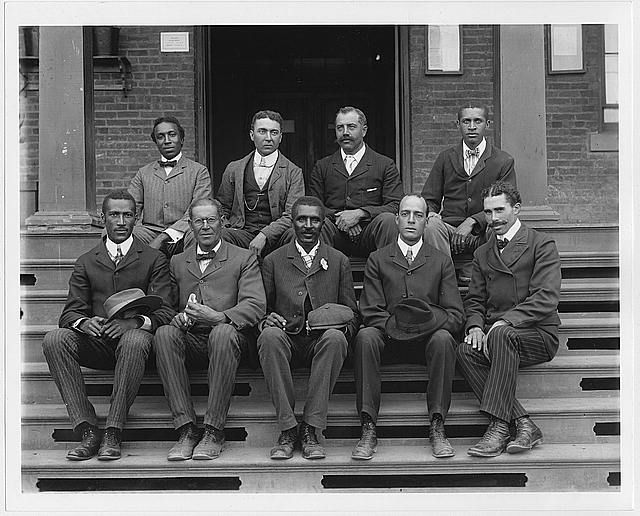 A black and white group photo with Dr. George Washington Carver seated on steps in the first row and in the middle with other staff at what is today known as Iowa State University. 