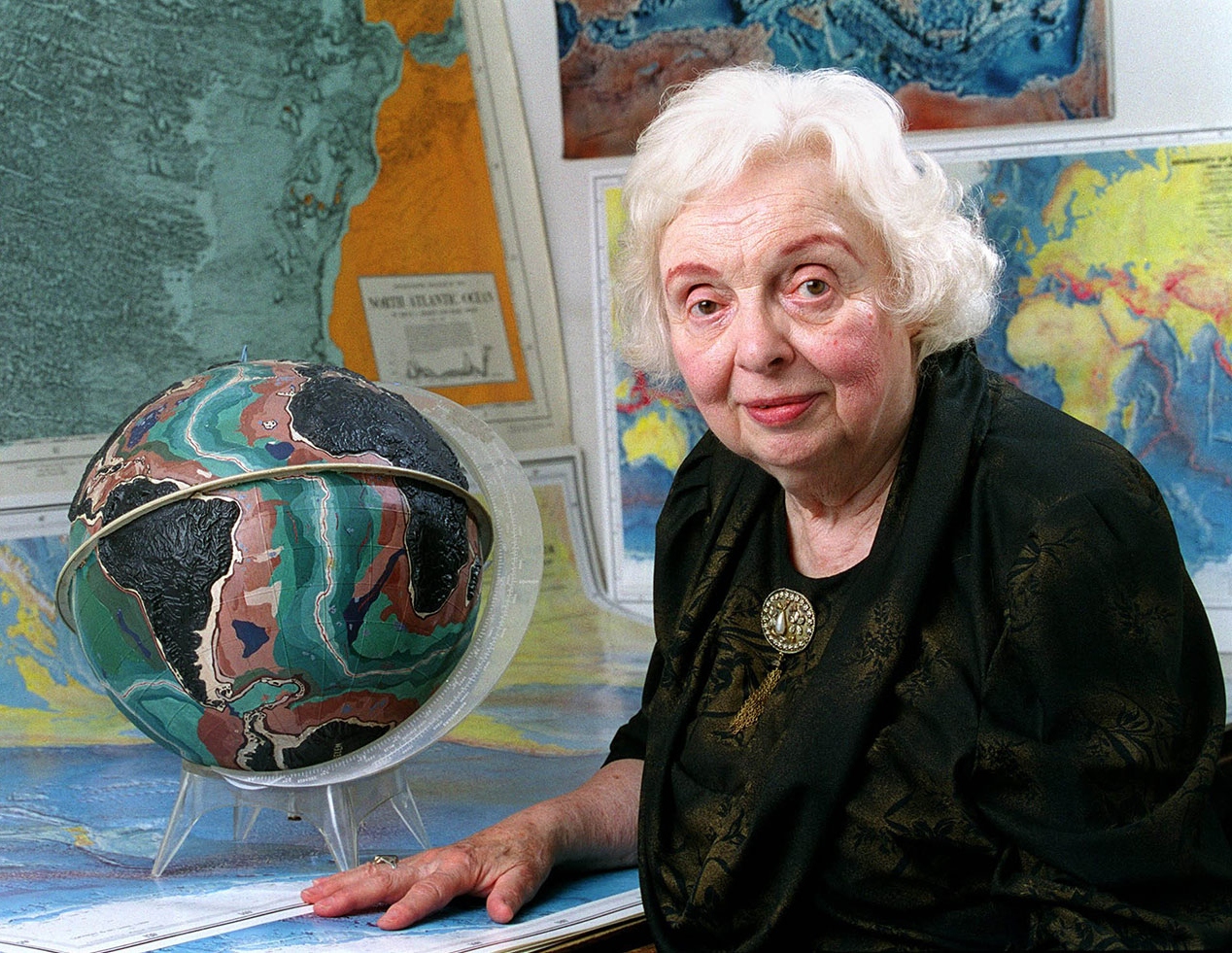 Marie Tharp standing next to a globe and in front of world maps in July 2001.