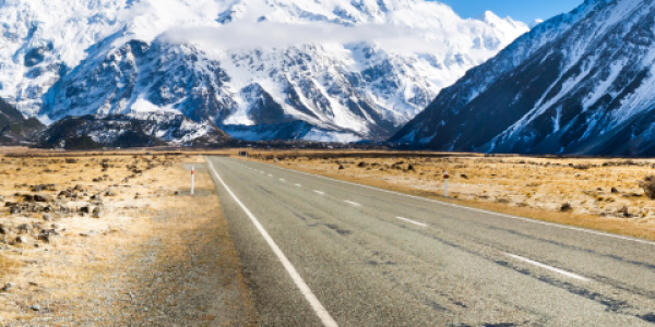 Paved road winding through New Zealand landscape with snow-capped mountains and cloudy sky in the background.