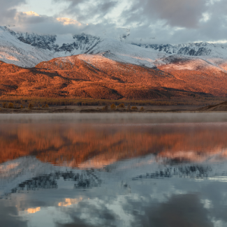 Snow capped mountains and sun turning the lower tier red in the light; mountains reflected in the lake.