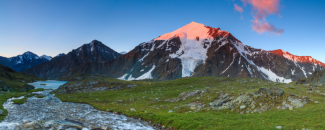 Snow-capped mountain in the background with a stream flowing over rocks to the left, and the sunset bathing the top of the mountain in red light.
