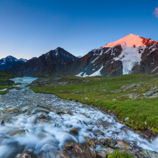 Snow-capped mountain in the background with a stream flowing over rocks to the left, and the sunset bathing the top of the mountain in red light.