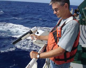 NCEI scientist Barry Eakins holds equipment as he stands at the rail of a ship in the ocean.