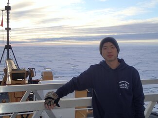 Scientist Elliot Lim standing on the deck of a ship in the Arctic with ice-covered water and a sunny sky behind him.
