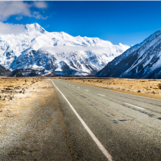 Paved road winding through New Zealand landscape with snow-capped mountains and cloudy sky in the background.