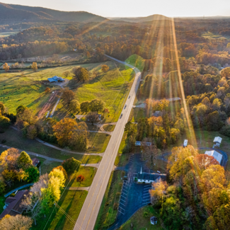 Aerial view of a backlit road in the Georgia mountains with green fields and trees that are beginning to turn to fall colors.