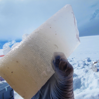 Gloved hand holding a piece of a cylindrical ice core from Hunter, AK outside in the snow. The ice has some small debris and is fairly cloudy at the bottom. Photo by Mike Waszkiewicz.