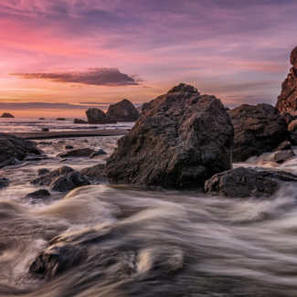 Rocky shoreline in the Pacific Northwest of the U.S., with the sun setting in the background, and turbulent water in the forefront of the image.